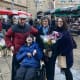 A smiling young man with cerebral palsy is seated in his wheelchair surrounded by his family in a town plaza market.